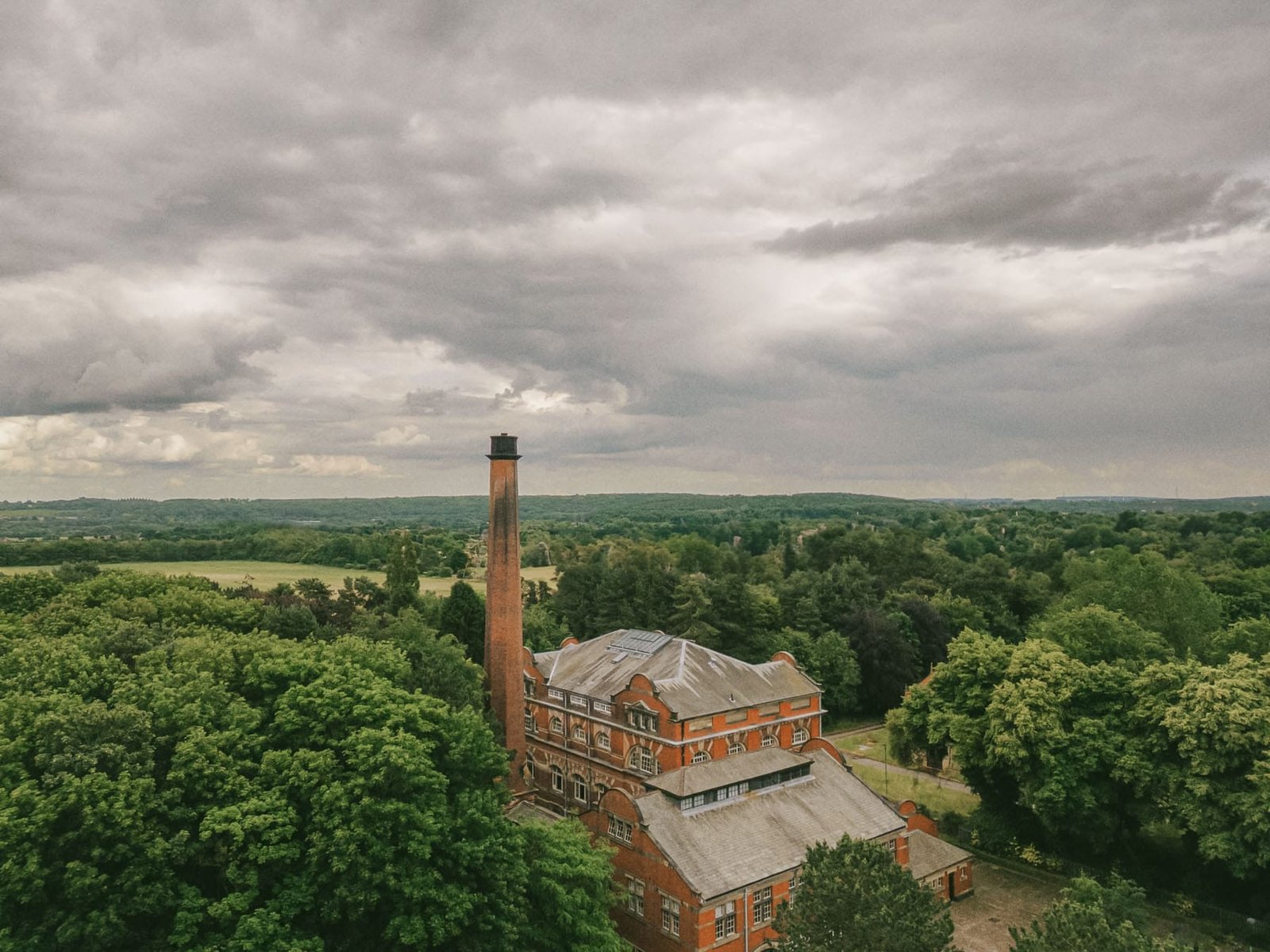 Aerial view of a red brick industrial building, reminiscent of a historic pumping house, with a tall chimney surrounded by lush green trees. Captured by drone, the landscape features rolling hills under an overcast sky with thick gray clouds, offering inspiration for any wedding photographer.