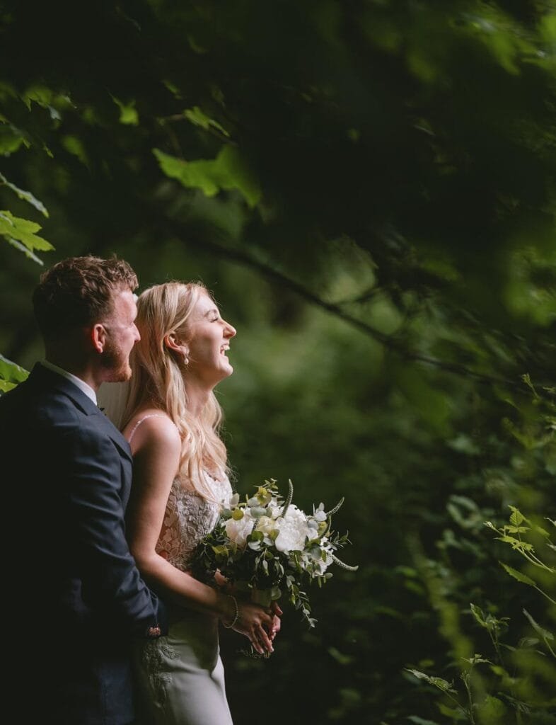 In the lush, green setting of The Pumping House wedding venue, a couple stands closely. The woman, holding a bouquet of white flowers, smiles brightly at the horizon. The man, wearing a suit, stands slightly behind her, sharing her joy as sunlight filters through the leaves.