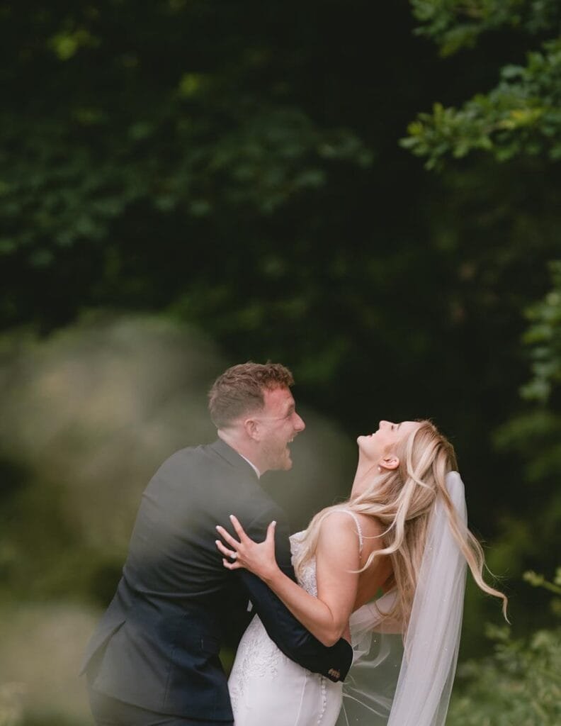 A bride and groom laugh joyfully amidst lush greenery at the Pumping House wedding venue. The bride, in a white gown and veil, leans back as the groom, in a black suit, holds her. They stand surrounded by blurred foliage, capturing a candid and happy moment.