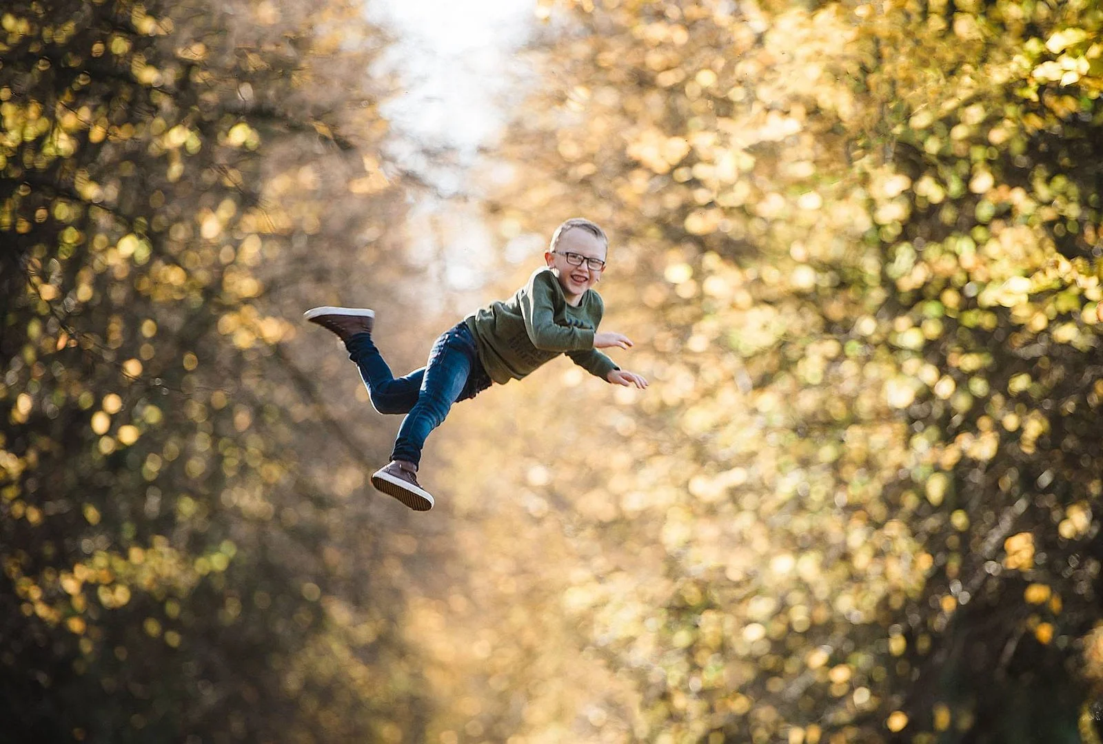 A young boy with glasses and a green jacket is flying through the air with a joyful expression in a park with autumn foliage.
