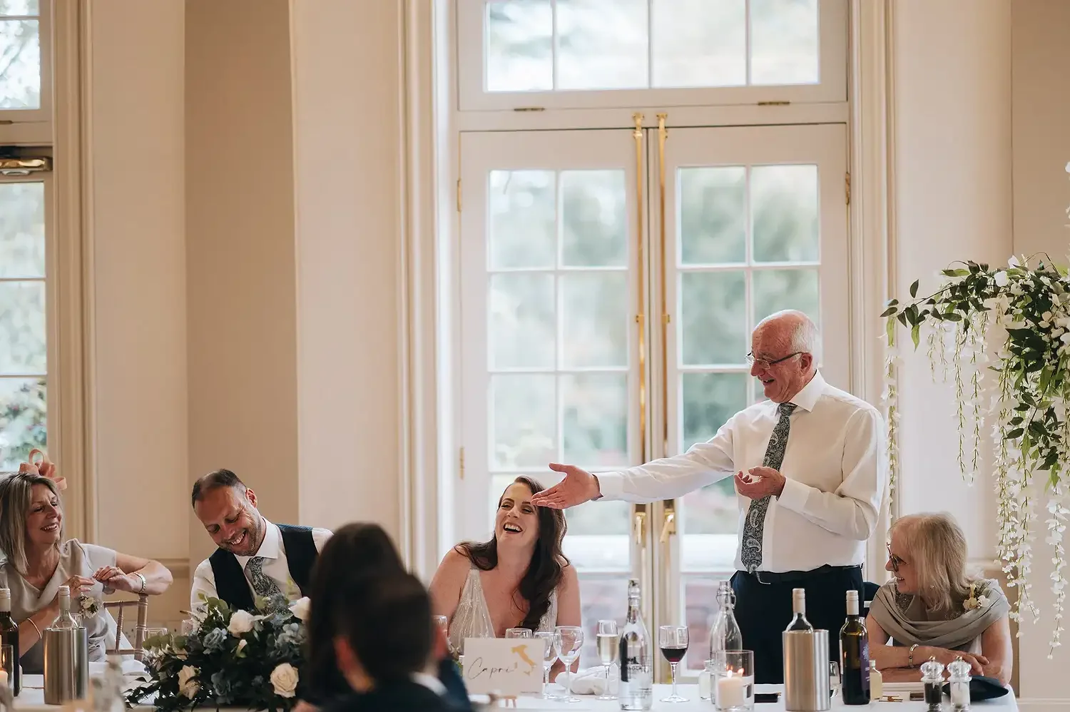 An elderly man giving a toast or speech at a wedding reception, with guests laughing and smiling around him in a decorated venue with large windows and natural light. Take at Hodsock Priory