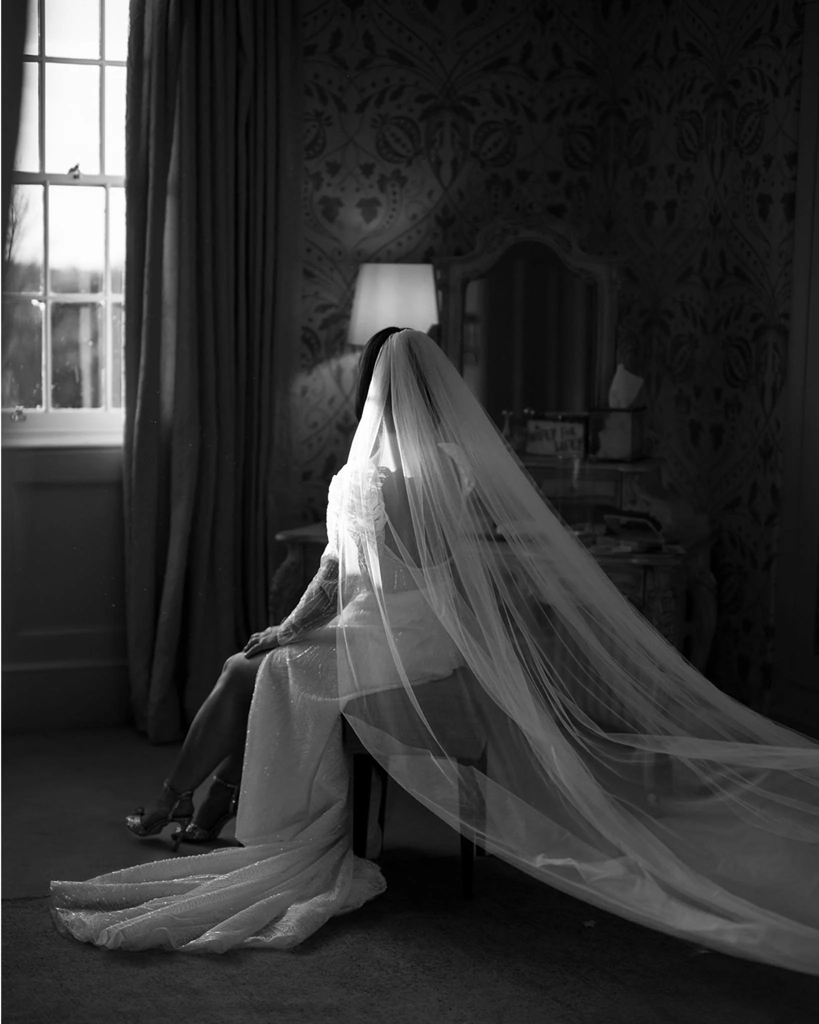A bride in a wedding dress and veil sitting on a chair in a dimly lit room next to a window with curtains, with a lamp and dresser in the background. Photograph taken inside of Hodsock Priory Bridal honeymoon suite