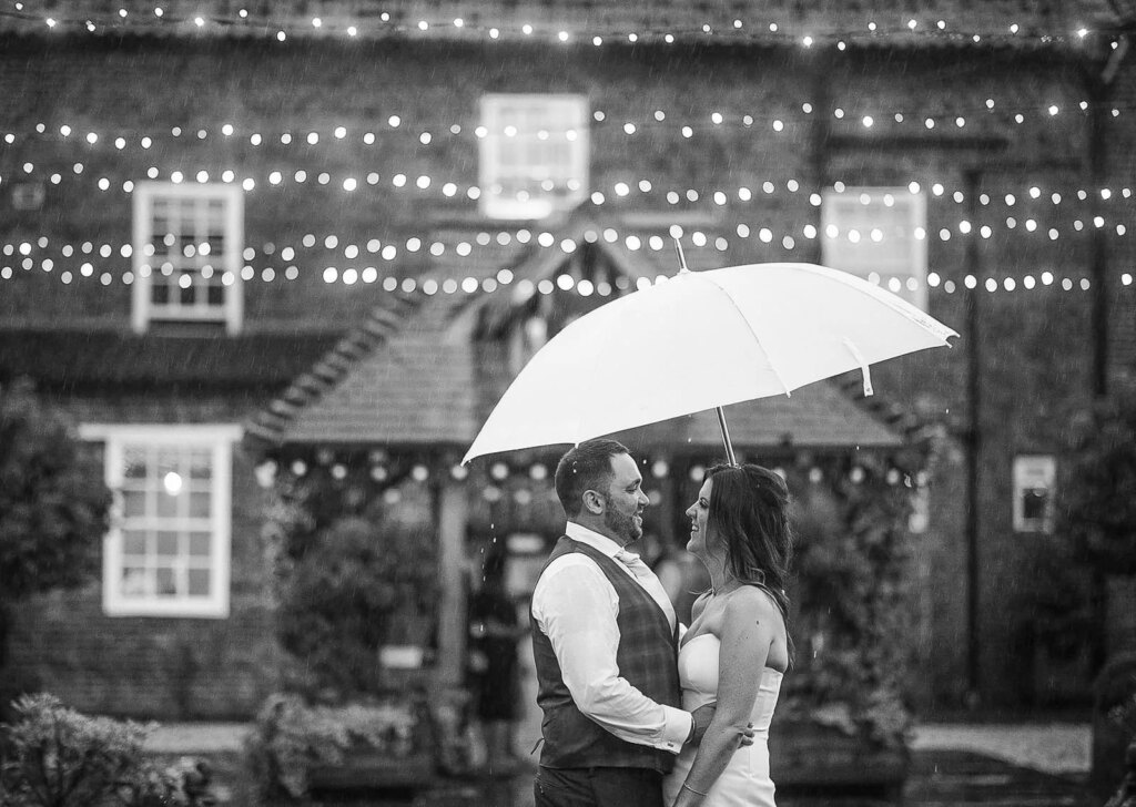 A couple in wedding attire is standing close together under an umbrella, sharing a joyful moment in the rain at night. String lights are hanging above them in front of a rustic brick building with windows and greenery.