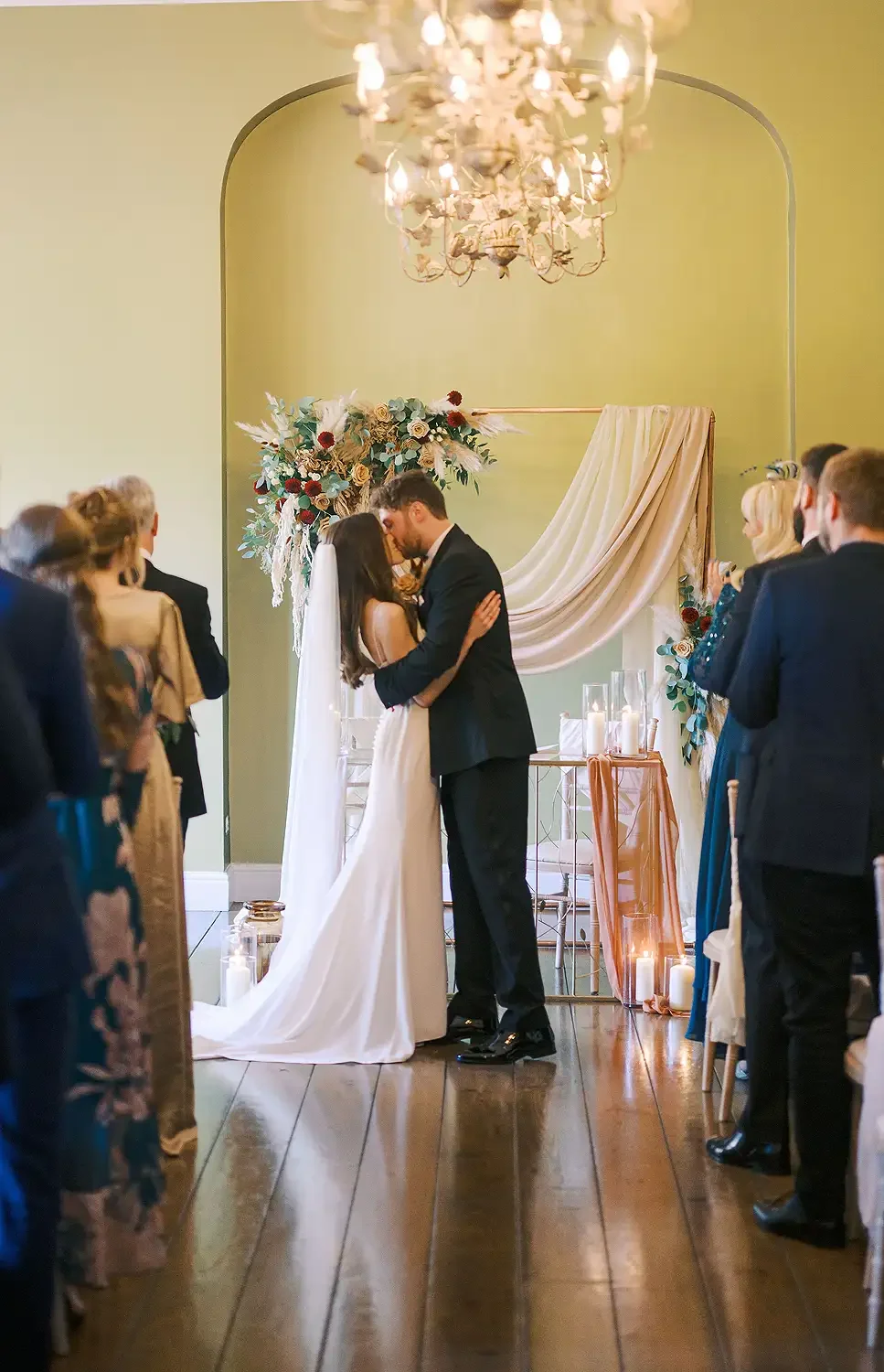 A wedding ceremony with a bride and groom sharing a kiss at the altar, surrounded by guests indoors with candlelit decor and floral arrangements. photograph of Hodsock Priory