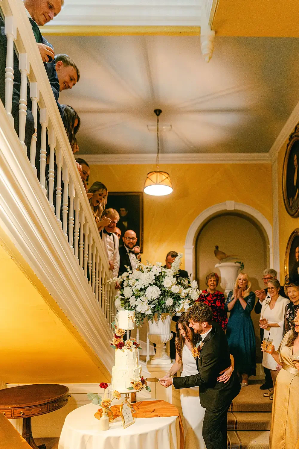 A wedding reception with the bride and groom cutting their wedding cake in a decorated elegant room filled with guests, flowers, and framed portraits. Take at Hodsock Priory