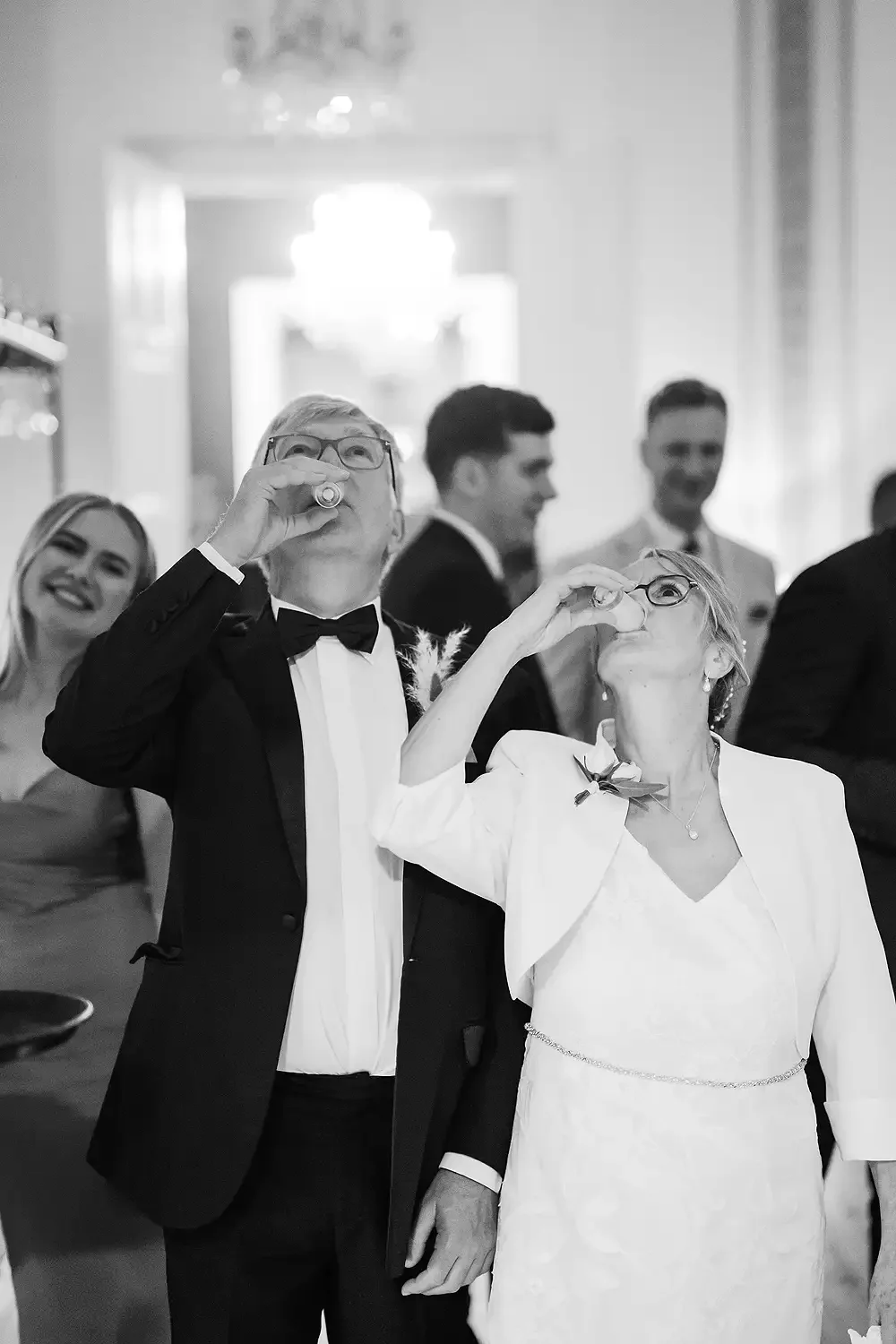 A group of people at a formal event, two of whom are drinking shots. The man is wearing a tuxedo and the woman a white dress with a floral accessory, both with glasses. There are smiling people in the background. Take at Hodsock Priory