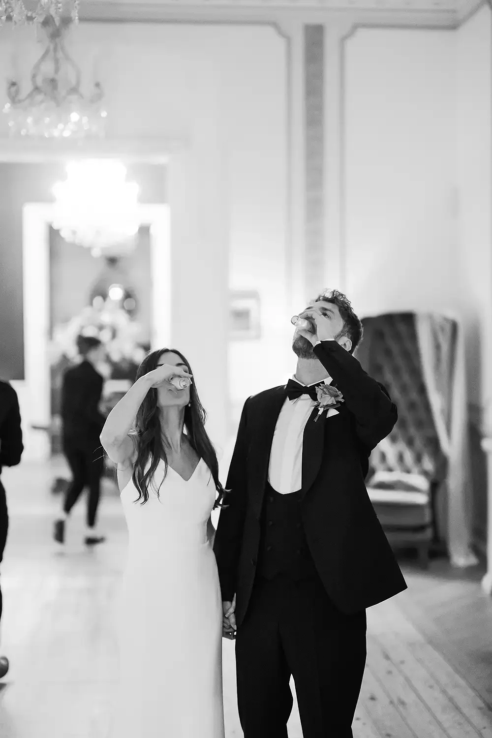 A bride and groom in wedding attire raising glasses for a toast at their wedding reception, holding hands. Taken at Hodsock Priory