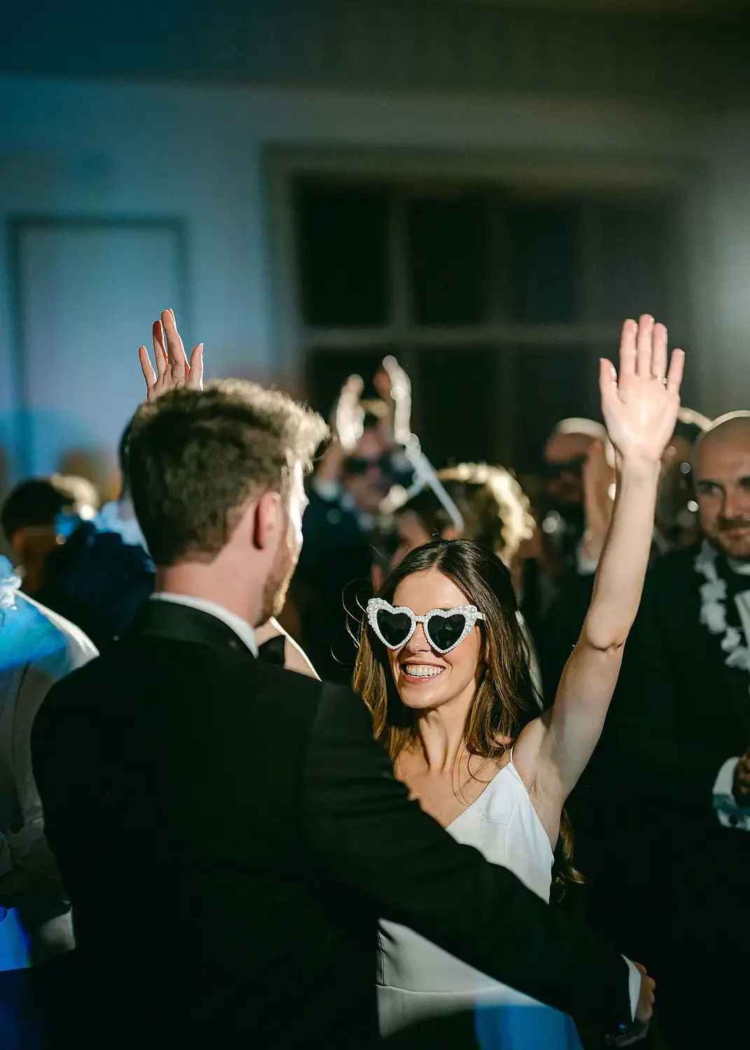 People dancing at a party or celebration, woman with heart-shaped sunglasses smiling and raising her arm, man in tuxedo looking at her. Taken at Hodsock Priory