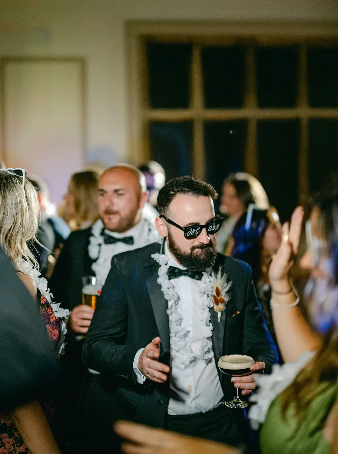 Man with a beard, sunglasses, and a flower boutonniere, wearing a tuxedo, holding a cocktail, at a social gathering or party. Taken at Hodsock Priory