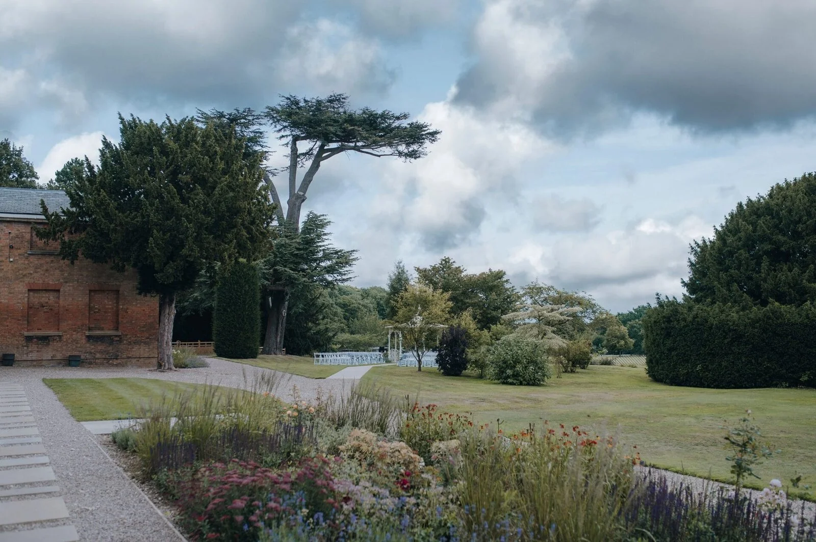 A scenic garden landscape with a brick building on the left, green trees and shrubs, a grassy lawn, a pathway, and a partly cloudy sky overhead. Take at Norwood Park