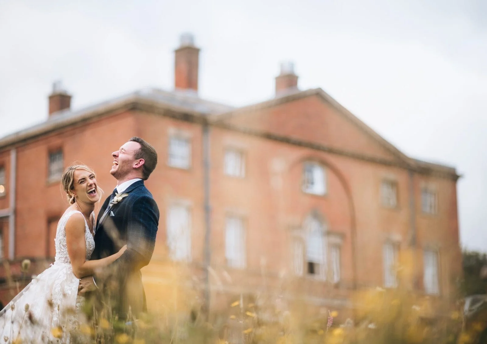 A bride and groom laughing and embracing outdoors in front of a large brick house, with grass and yellow flowers in the foreground. Take at Norwood park