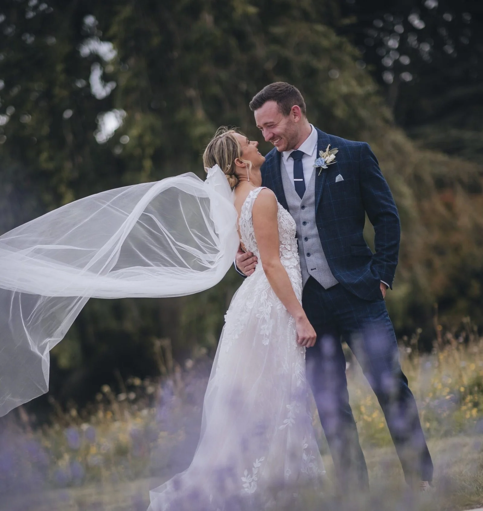 A bride and groom on their wedding day outdoors, standing close and smiling at each other with the bride's veil flowing behind her. Take in the gardens of Norwood Park