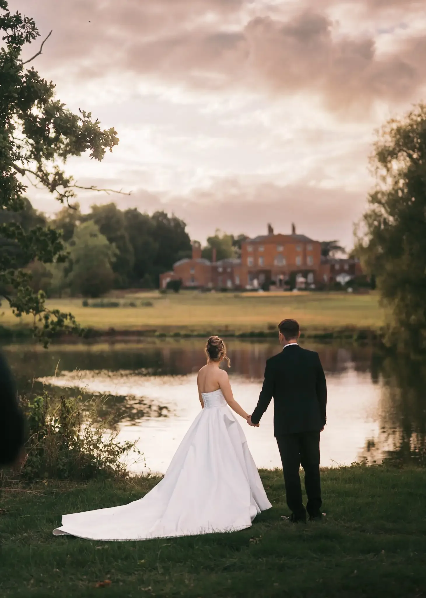 A bride in a white wedding gown and a groom in a black suit holding hands by a lake at sunset, with a large house in the background. Norwood Park on a wedding day in the bakground