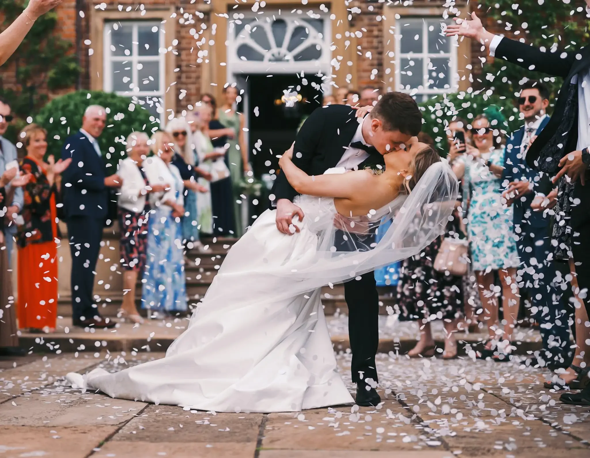 A newlywed couple sharing a kiss as guests throw confetti around them outdoors in front of a brick building with white windows and a door, in the grounds of Norwood Park