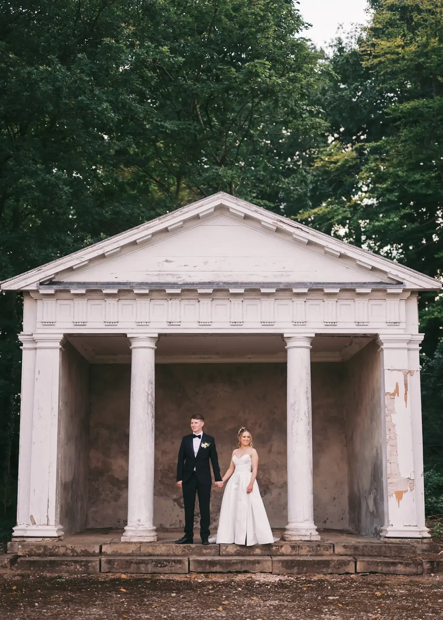 A bride and groom standing hand in hand in front of a white, classical-style Temple with tall columns, greenery in the background, and a weathered appearance, in the grounds of Norwood Park