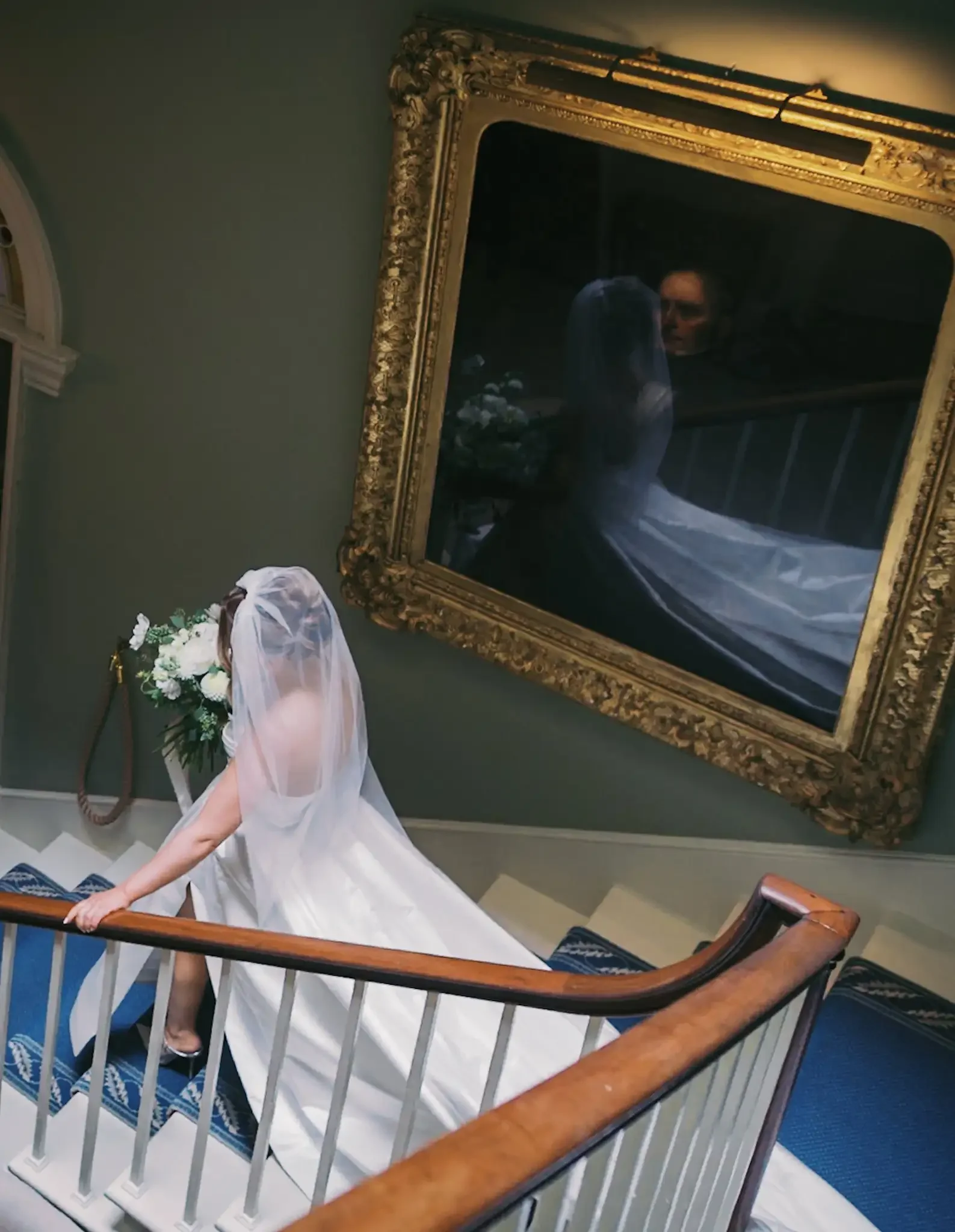 A bride in a white wedding dress with a veil walking up a staircase with a floral bouquet in her hand, photographed from the side. A large ornate gold-framed mirror is on the wall behind her, reflecting her and the staircase of Norwood Park