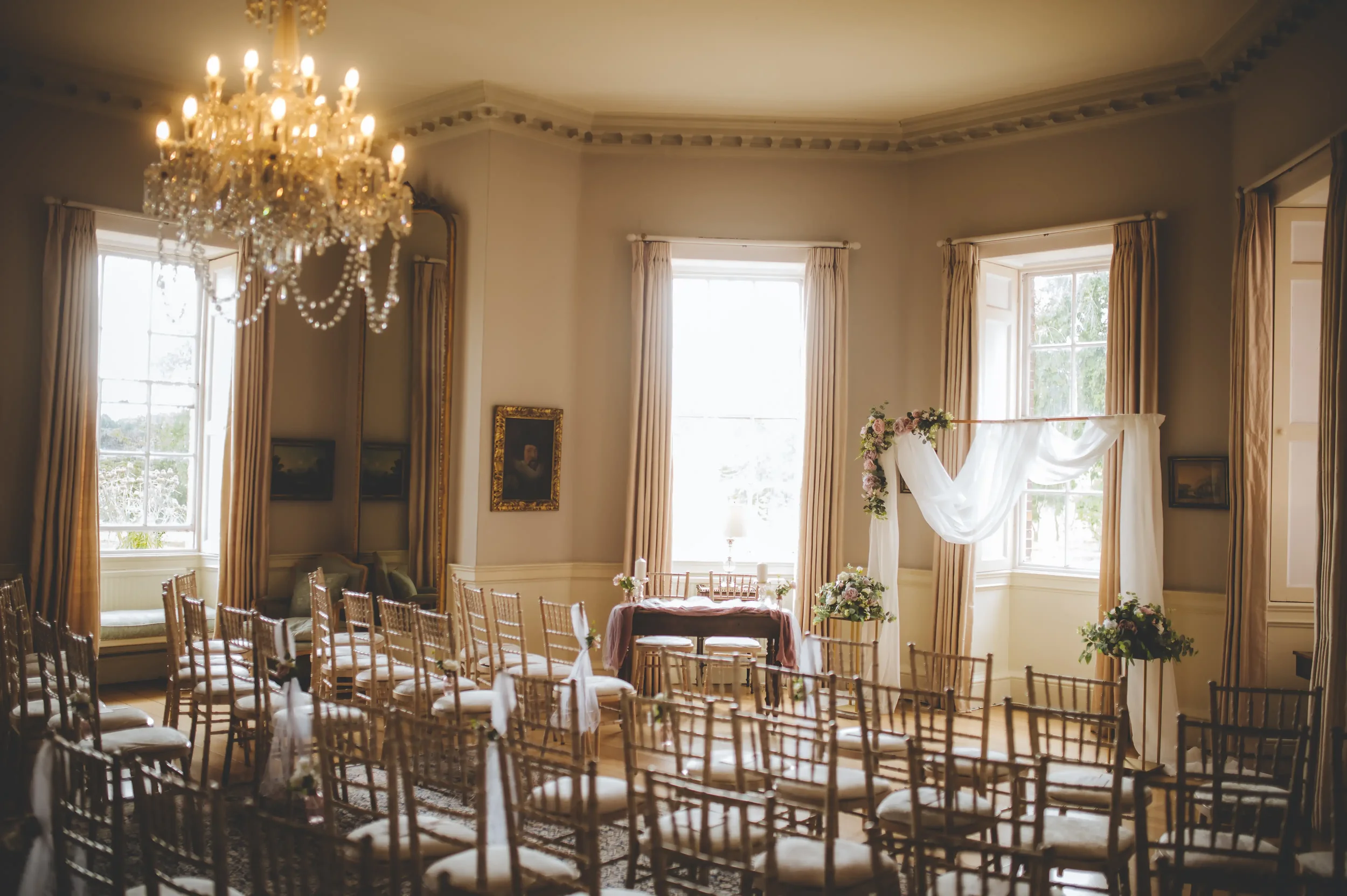 Elegant wedding ceremony setup in a bright, vintage-style room with a chandelier, gold chairs, floral arrangements, and a draped white fabric on a wooden arch near large windows. Take at Norwood Park