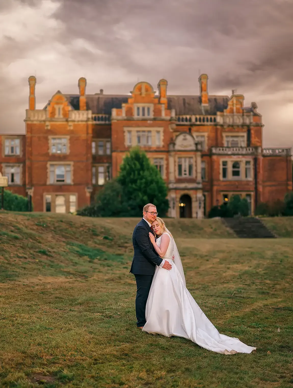 A bride and groom embrace on a grassy hill with a large historic brick building and a dramatic sky in the background. Take at Rossington Hall at sunset