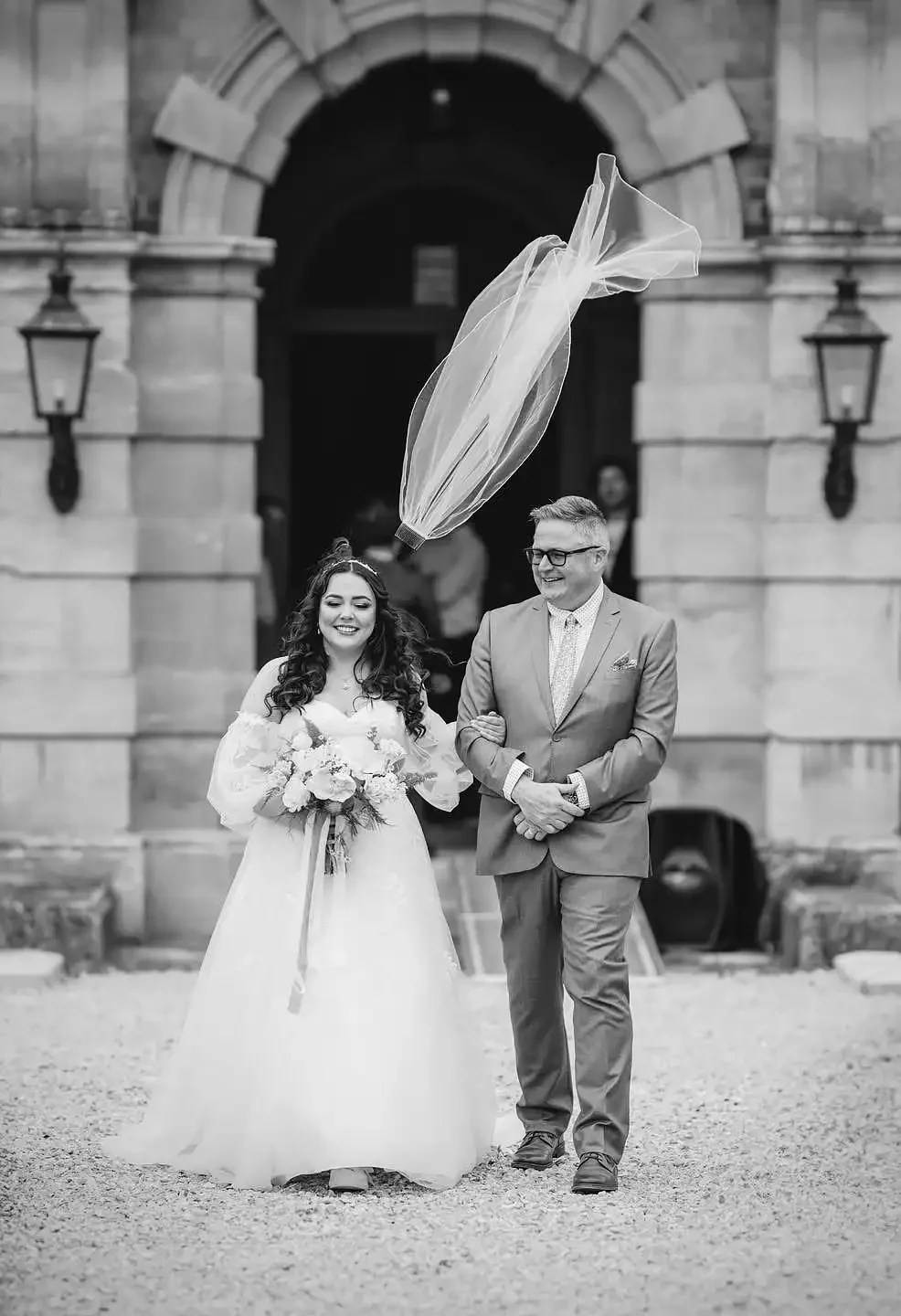 Black and white photo of a bride in a wedding dress holding a bouquet, walking arm-in-arm with a smiling groom in a suit, outside a historic building with arched doorway, with a large balloon floating above them. Taken at Rossington Hall