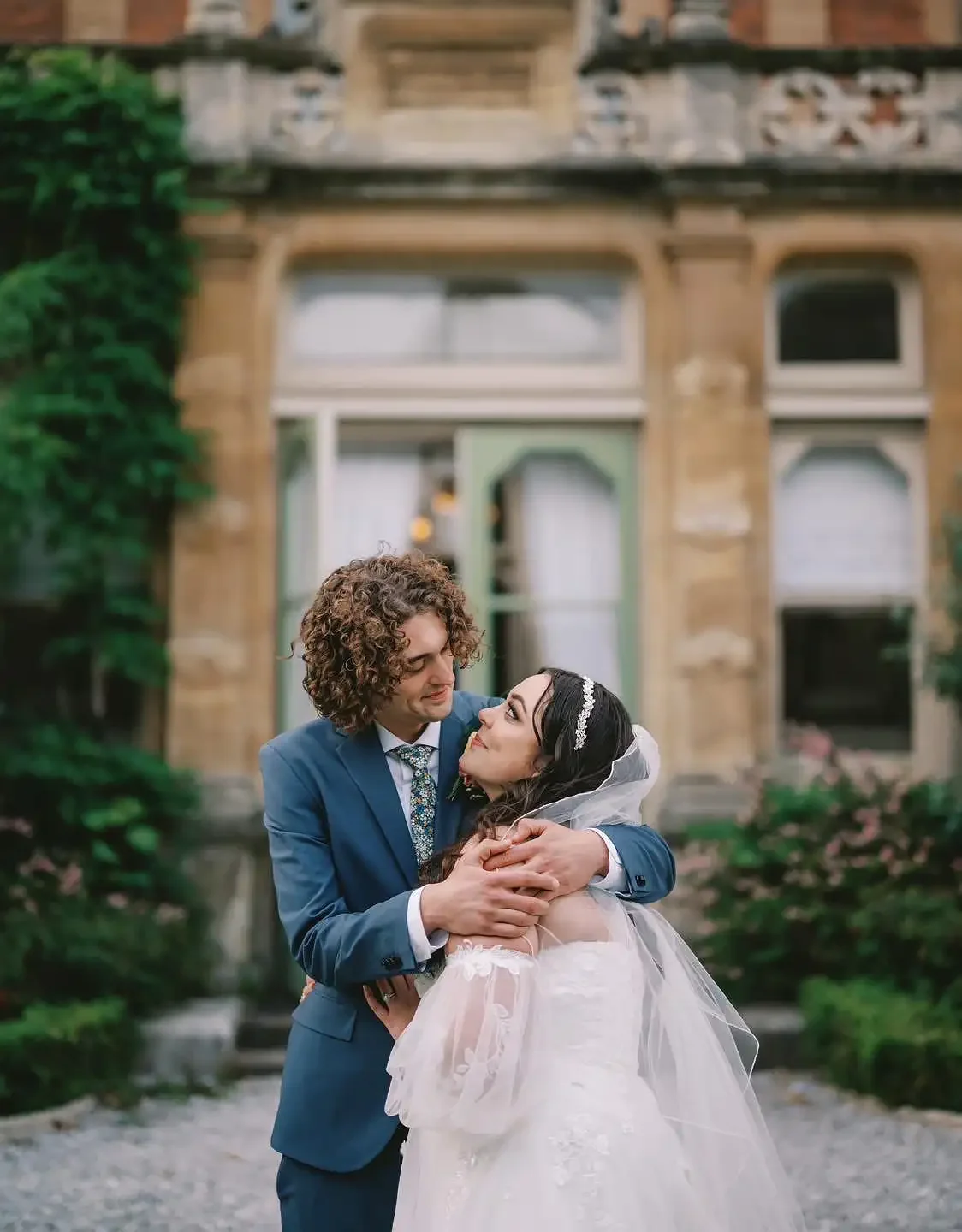 A bride and groom embrace outdoors in front of a historic building with large windows and stone architecture, greenery, and pink flowers. Take at Rossington Hall