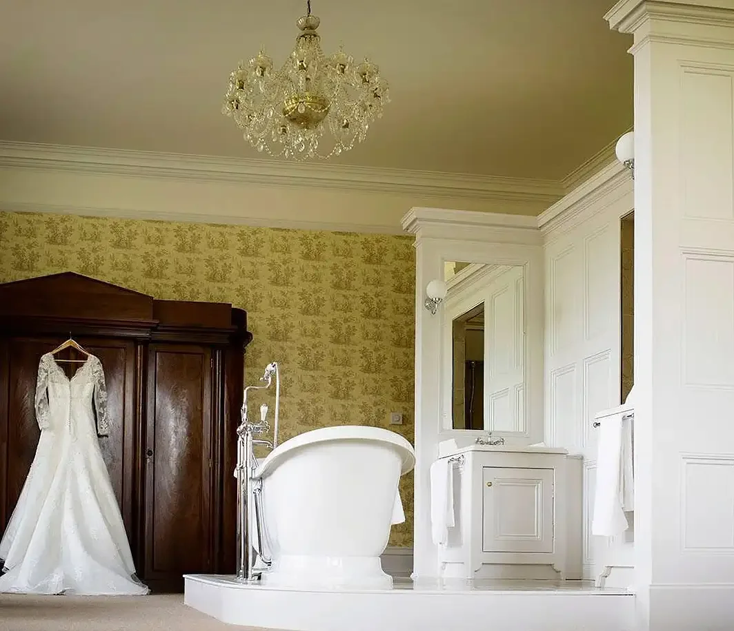 A wedding dress hanging inside a wooden wardrobe near a vintage bathtub in a luxurious bathroom with a chandelier and yellow wallpaper. taken in the honeymoon bridal suite of rossington hall