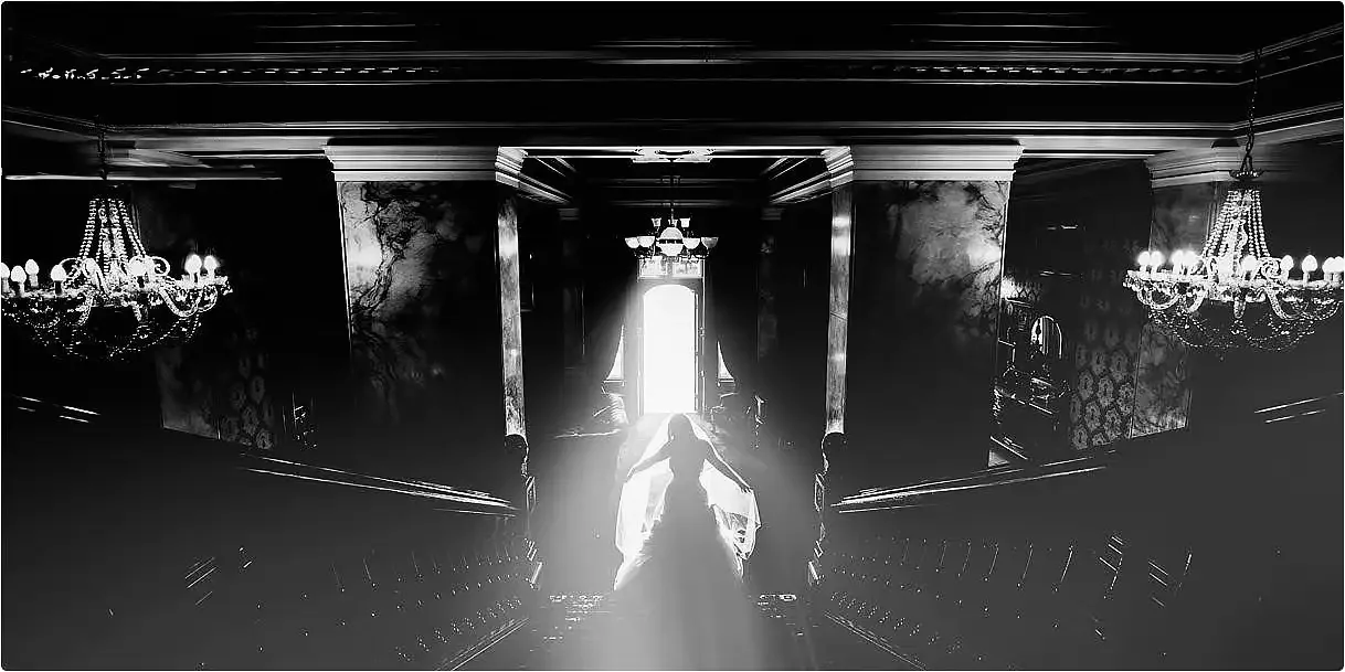 Silhouette of a person in a gown walking in a grand, vintage mansion interior with chandeliers and a bright door at the end of the hallway. Take at Rossington Hall from the staircase