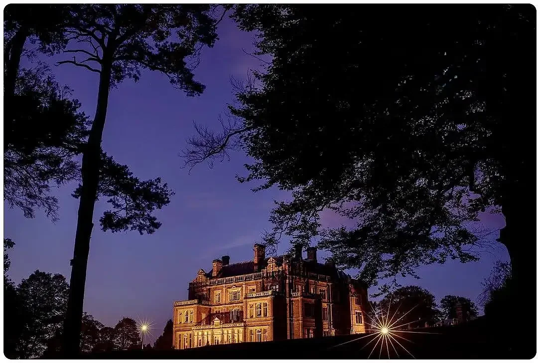 A large, historic mansion illuminated at night, framed by silhouettes of trees against a deep blue sky. Outdoor photo at a wedding at Rossington Hall