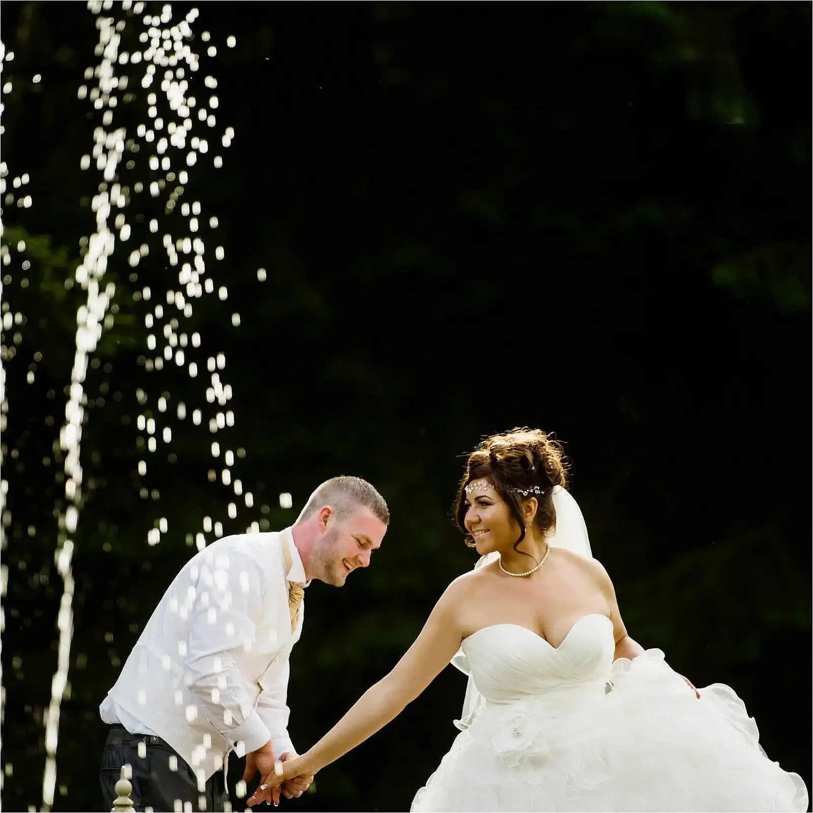 A bride and groom holding hands and smiling outdoors during their wedding, with water splashing in the air and a dark background. in the sunken garden of Rossington Hall