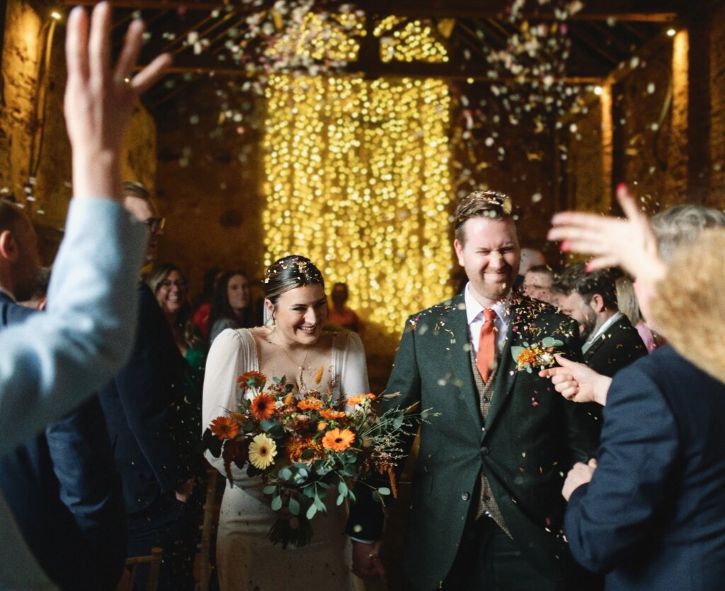 A bride and groom at their wedding celebration, surrounded by guests throwing confetti and smiling. Take in the barn at The Normans wedding venue york