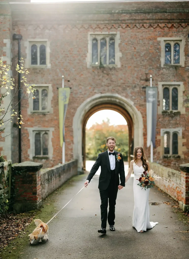 Bride and groom laughing as they walk out of the Hodsock Priory gatehouse with their dog on a lead