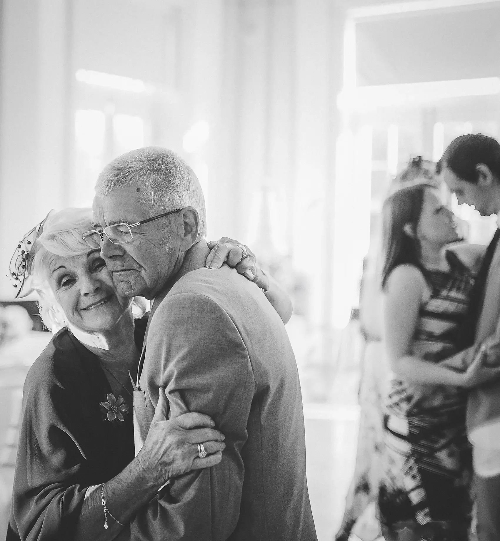 An elderly couple dancing closely at a celebration, with the woman smiling and the man wearing glasses, in a bright room with large windows. Taken at Hodsock Priory