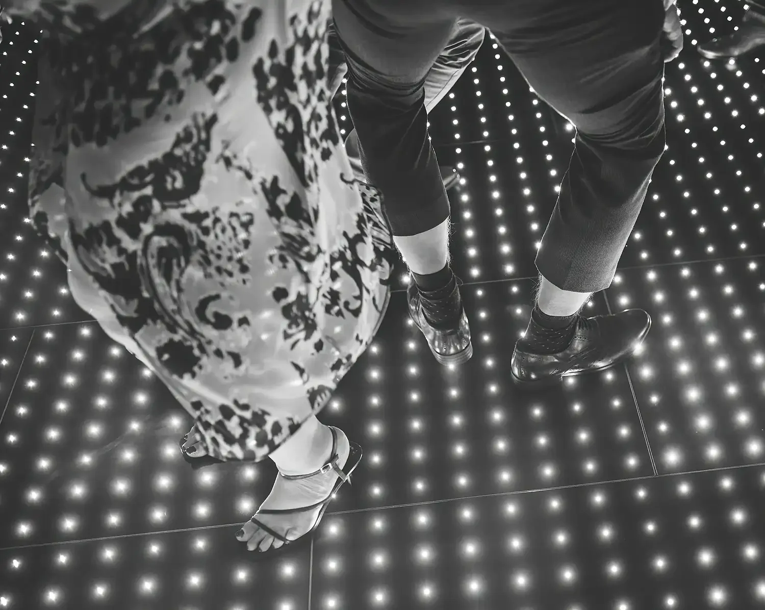 Black and white photo of two people standing on a floor with LED lights, showing their legs and feet, with one person wearing sandals and the other wearing dress shoes. Taken at Hodsock Priory