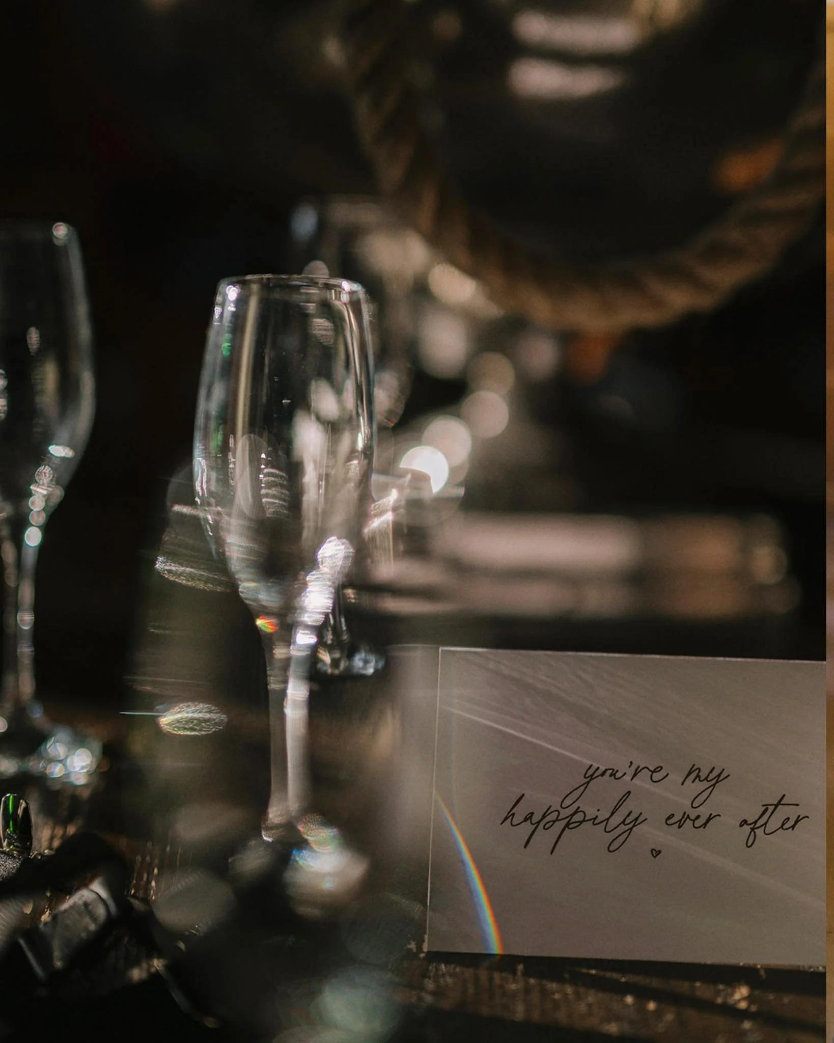 A close-up of empty champagne glasses on a table with a romantic note that reads "you're my happily ever after" and a rainbow reflection, suggesting a celebration or special occasion. Take at Hodsock Priory