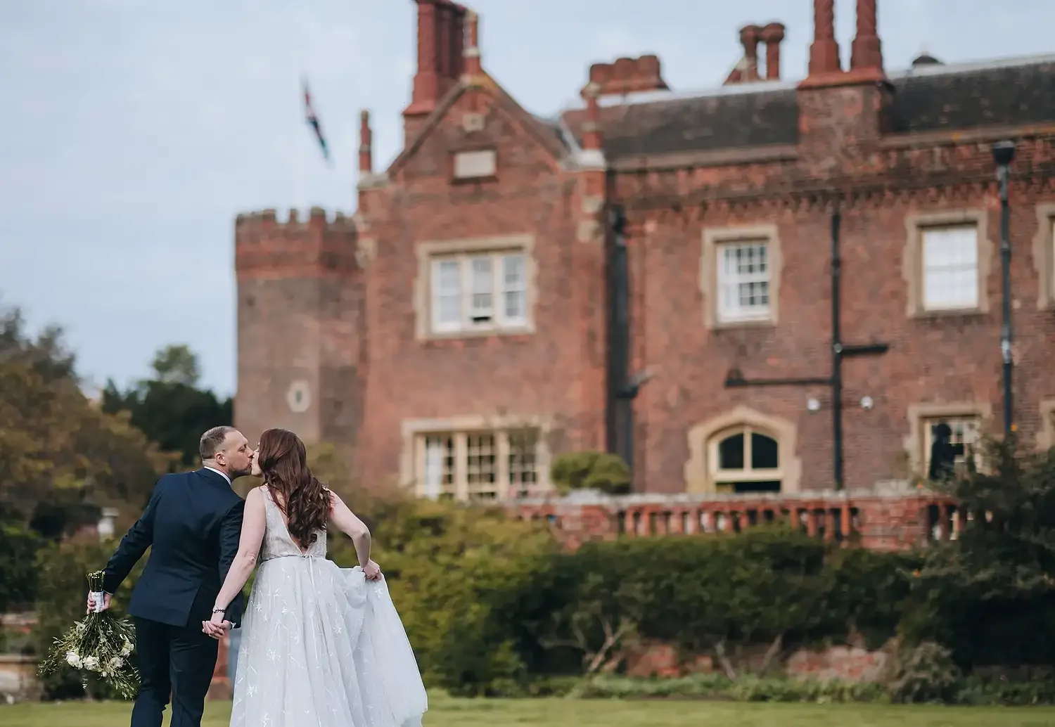 A bride and groom kissing in front of a large brick castle, with the bride holding a bouquet and the groom holding the bride's hand. Taken at Hodsock Priory