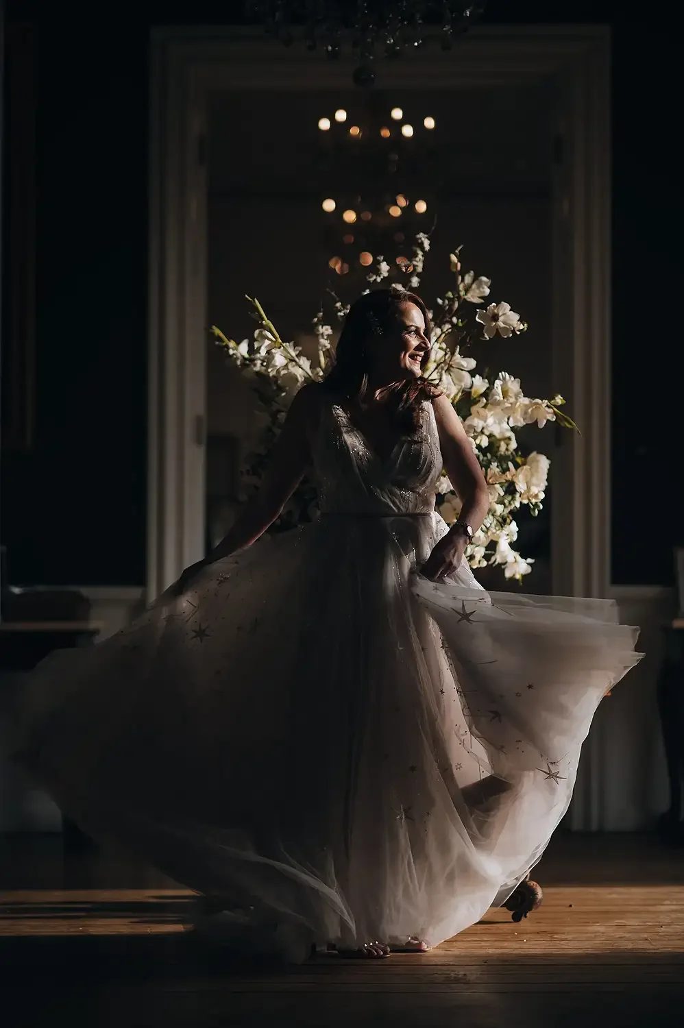 A woman in a light-colored, star-embellished dress dancing indoors with a large floral arrangement and chandelier in the background. Take at Hodsock Priory