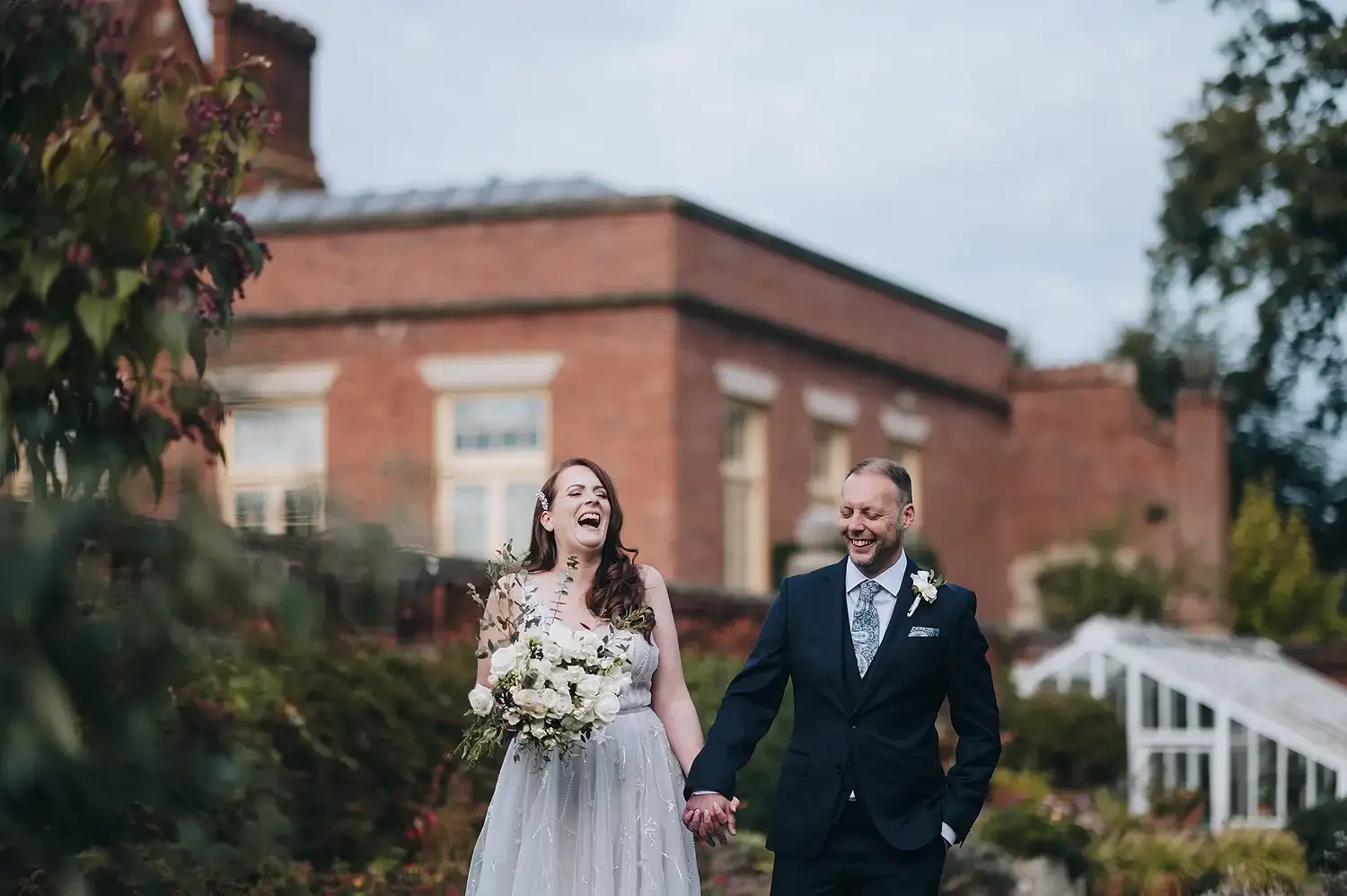 A bride and groom walking hand in hand outdoors, happily laughing, with a brick building in the background. Taken at Hodsock Priory