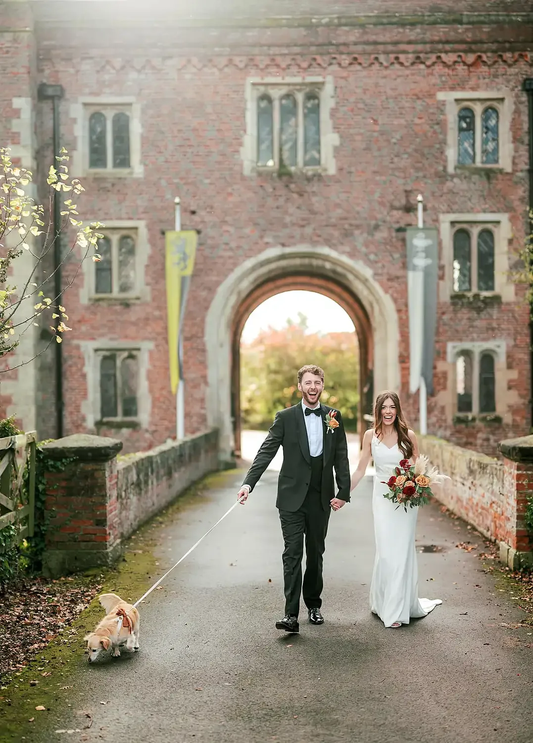 A newlywed couple walking outdoors on a paved path, smiling and holding hands; the groom in a tuxedo with a bow tie and boutonniere, and the bride in a white dress holding a bouquet of flowers; a small dog on a leash. Photograph of Hodsock Priory