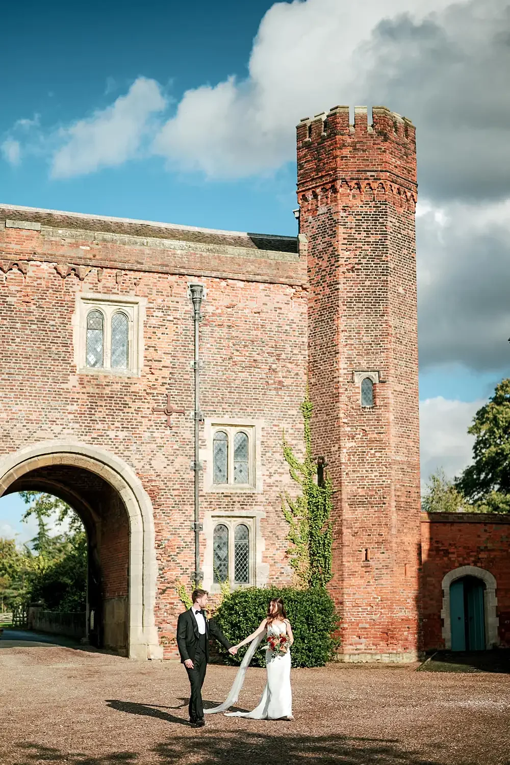 A bride and groom holding hands outside a historic brick building with a tall tower, lush green bushes, and cloudy sky. photograph of Hodsock Priory