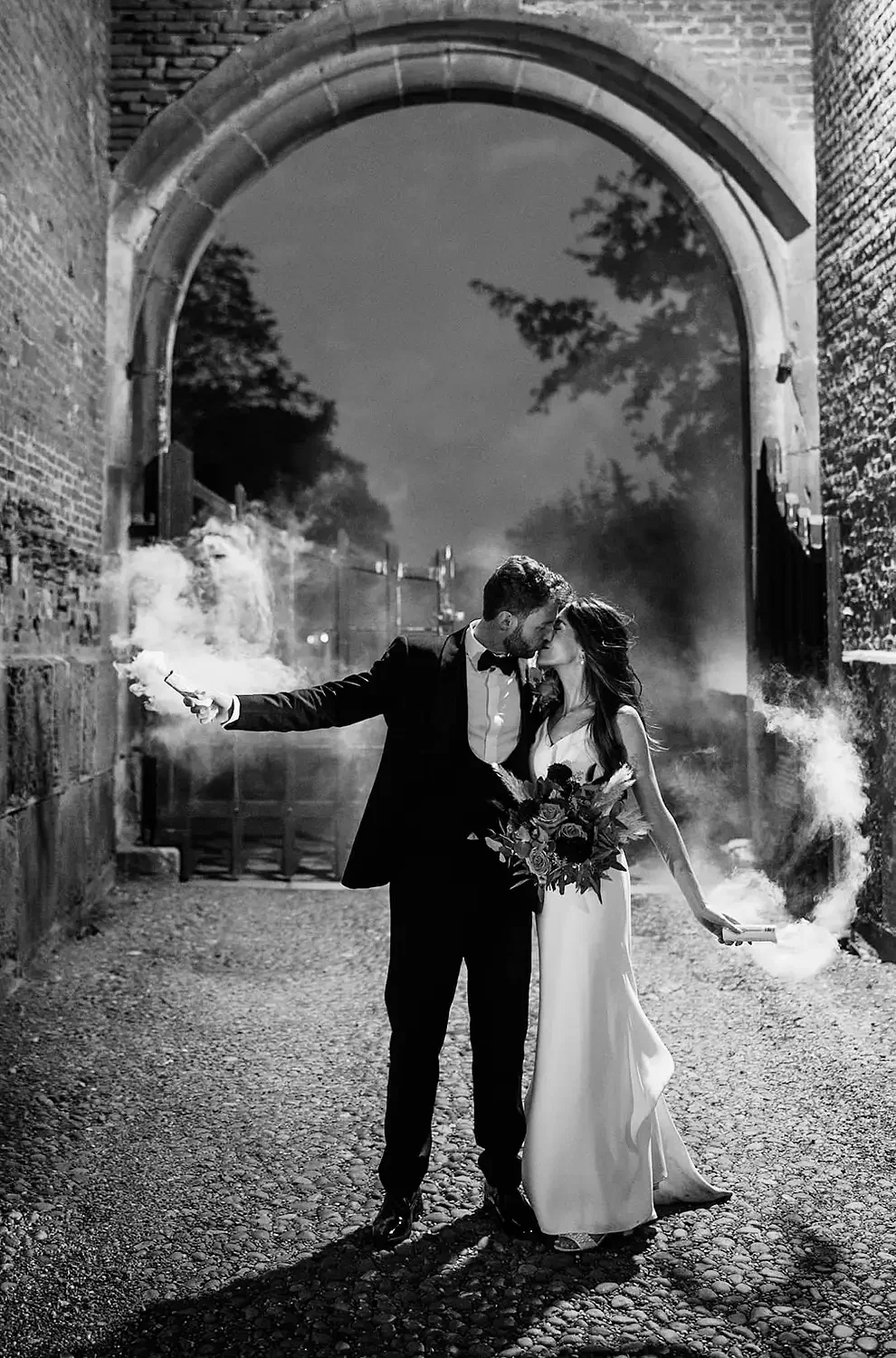 A black and white photo of a wedding couple standing under an arched gateway with smoke bombs in their hands, at Hodsock Priory. The bride is holding a bouquet and wearing a long dress, while the groom is dressed in a suit and bow tie.