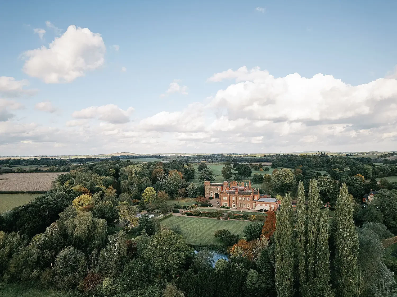 A large estate with a castle-like mansion surrounded by trees and greenery, under a partly cloudy sky. Drone photograph of Hodsock Priory