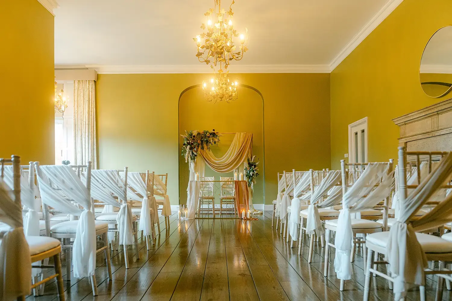 Elegant indoor wedding ceremony setup with white chairs draped in white fabric, a floral arch, and candles on the floor, in a room with yellow green walls, wooden floors, chandeliers, and a mirror. Buchanon Room Hodsock priory