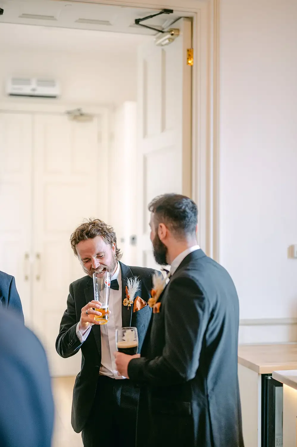 Two men in tuxedos, one with a beard and the other with curly hair, smiling and talking while holding drinks at a formal event or wedding reception. Take at Hodsock Priory