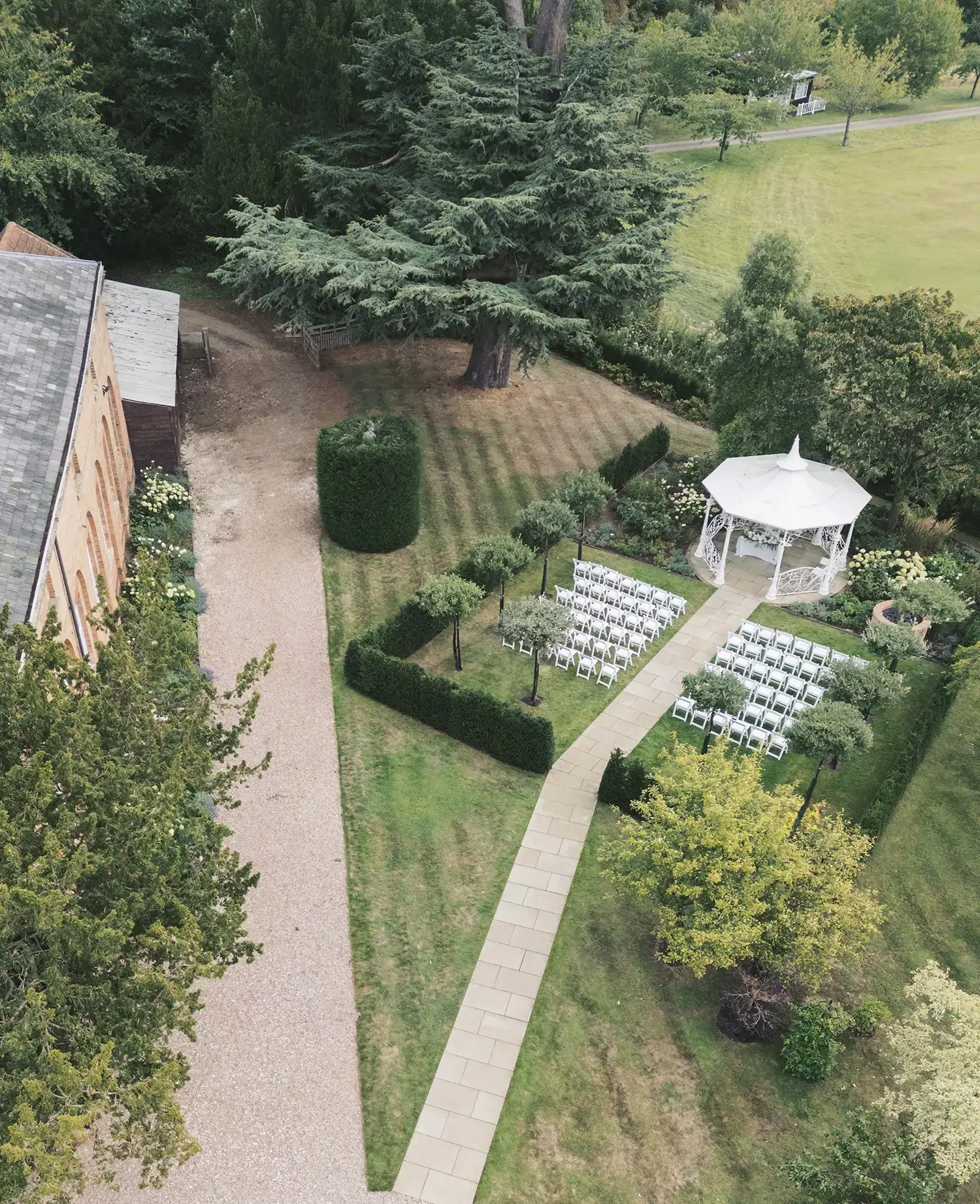 Aerial view of a garden wedding setup with white chairs arranged in rows, a white gazebo, small trees, bushes lining the walkway, a large tree, and a building on the left side. Drone shot of outdoor wedding at the Starkey Bandstand at Norwood Park