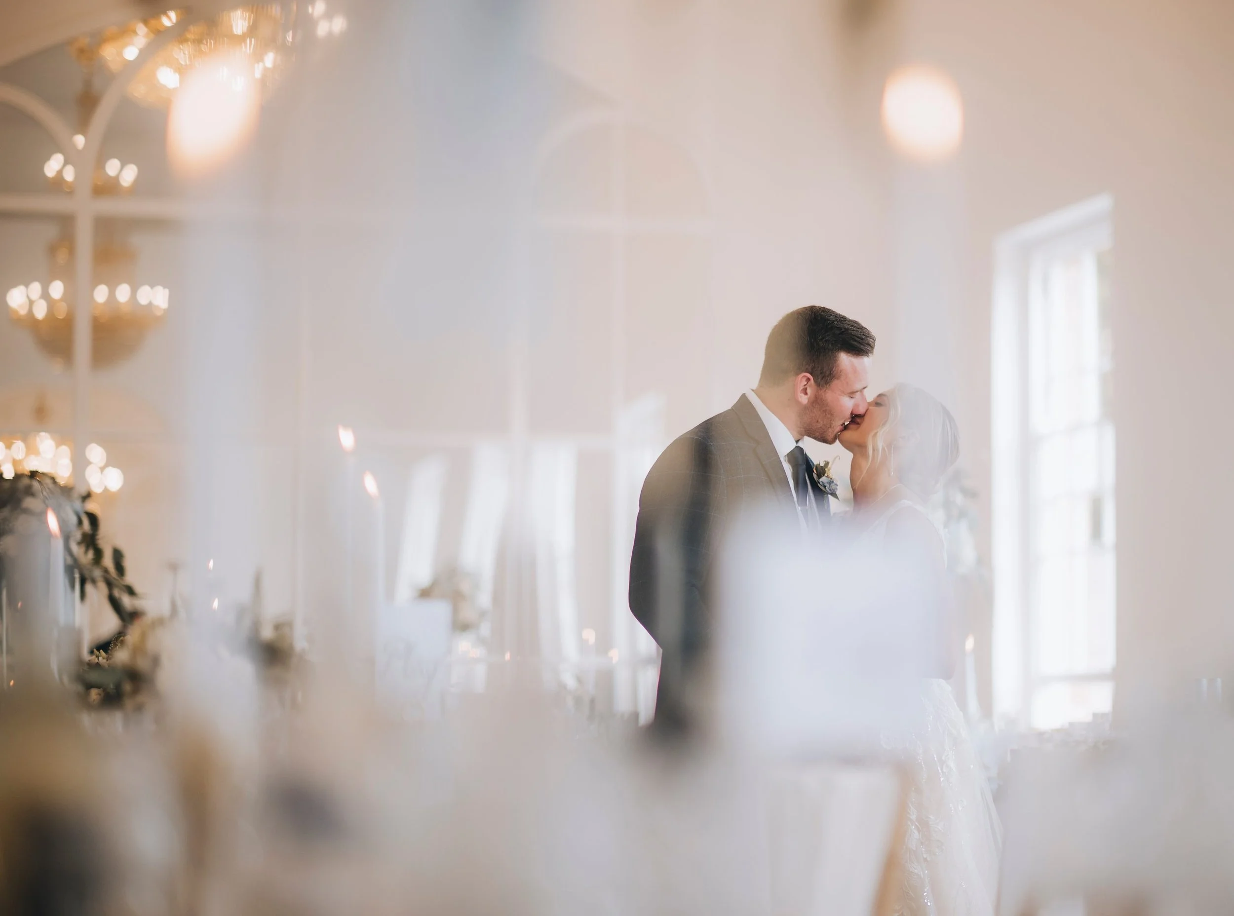 A newlywed couple sharing a kiss indoors, with soft natural lighting and elegant decor, seen through a blurred foreground that adds a romantic atmosphere. Take at Norwood Park