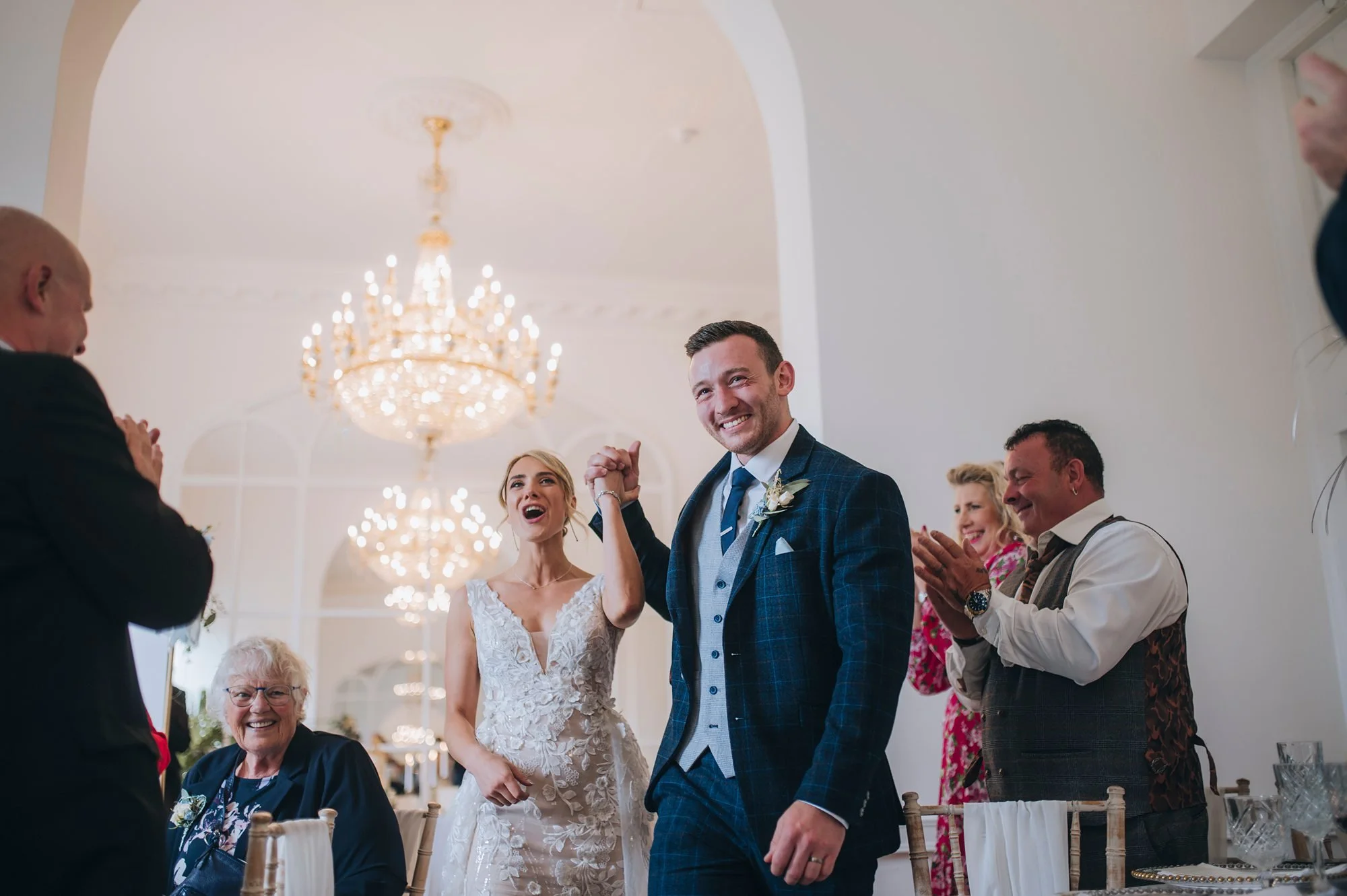 A wedding reception scene with a smiling groom holding the hand of a smiling bride, surrounded by clapping family members in a beautifully decorated venue with chandeliers. Take at Norwood Park