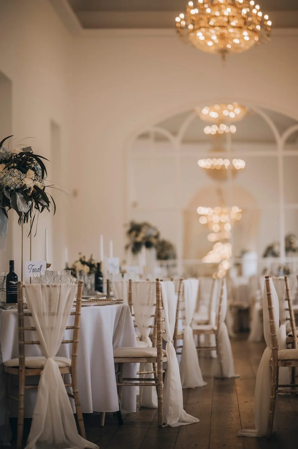 Elegant banquet hall decorated for a wedding with white tablecloths, beige chairs with tied sashes, floral centerpieces, and large chandeliers. Take at Norwood Park