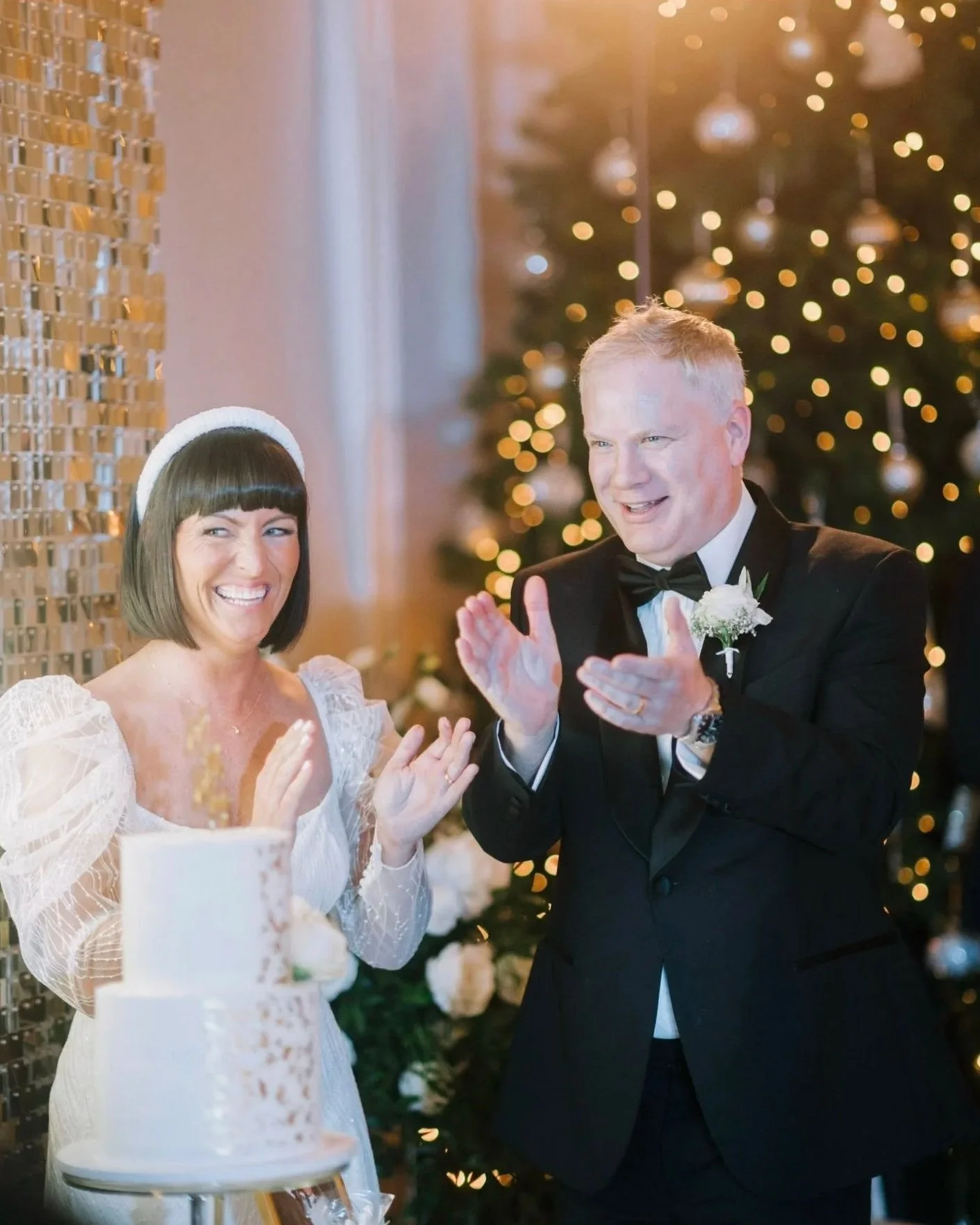 A bride and groom celebrating at their wedding with a decorated Christmas tree in the background. They are smiling and clapping near a wedding cake. Take at Hodsock Priory