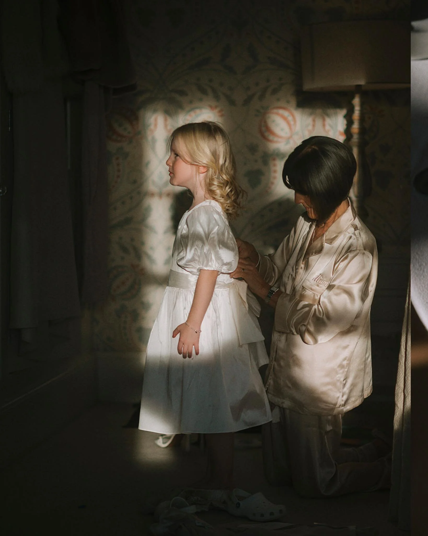 A young girl in a white dress standing while a woman, possibly her mother, helps her with her dress in a softly lit room with a patterned wallpaper in the background. Inside photograph of Hodsock Priory