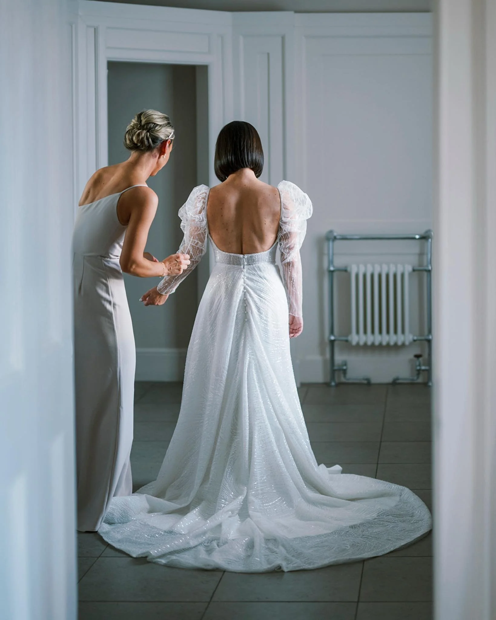 A bride in a white wedding gown with a low back and puffed sleeves is adjusting her dress while another woman in a long ivory dress helps her in a room with white paneled walls and a radiator. Photograph inside Hodsock Priory bridal suite