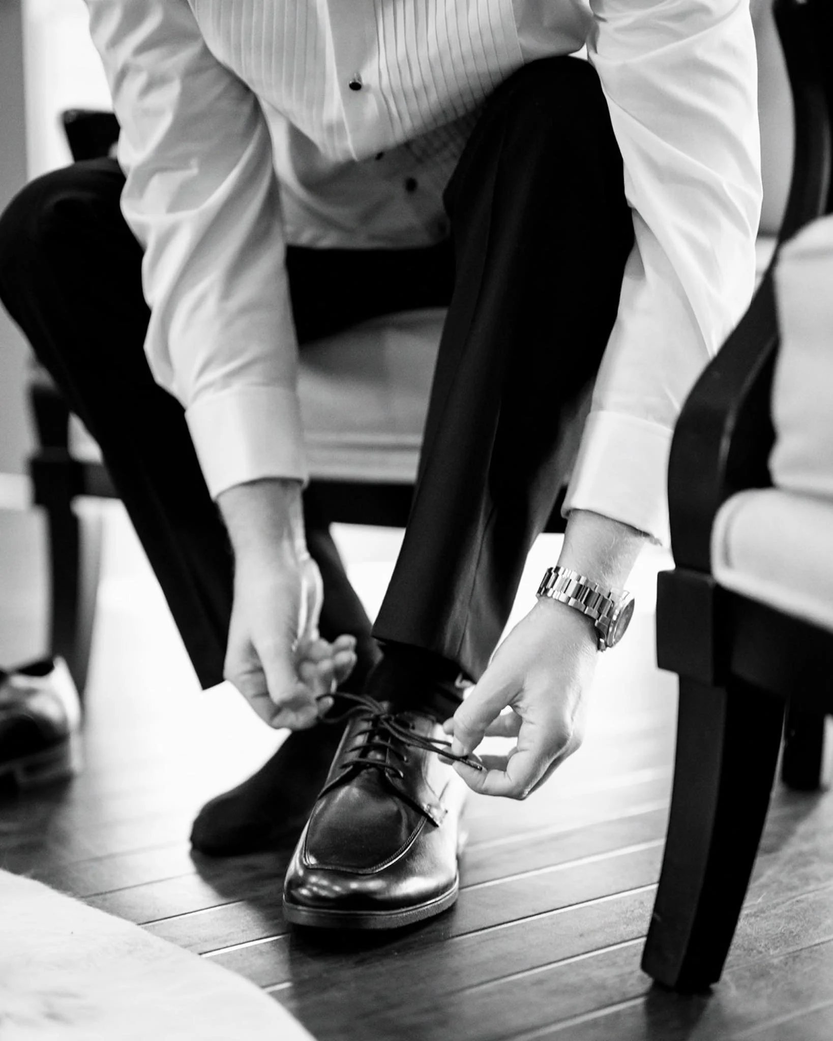 A man in formal attire sitting on a chair, tying his dress shoe. Take at Hodsock Priory